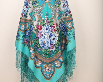 May include: A teal blue shawl with a floral pattern and fringe. The shawl is folded and displayed on a wooden mannequin.