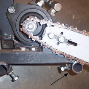 May include: Close-up of a metal chain saw sharpener with a black metal base and a silver chain saw blade. The chain saw blade is attached to a metal wheel with a black rubber belt. The chain saw sharpener has two metal bolts on the base.
