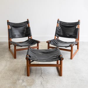 May include: Two black leather chairs with wooden frames and a matching black leather ottoman with a wooden frame.