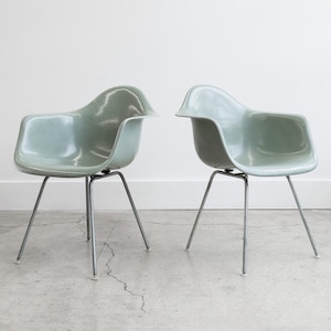 May include: Two light green molded plastic armchairs with metal legs. The chairs have a rounded seat and back, and are set against a white wall and gray floor. The chairs are a mid-century modern design.