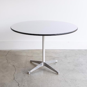 May include: A white round table with a black edge and a silver star-shaped base. The table is on a gray concrete floor.