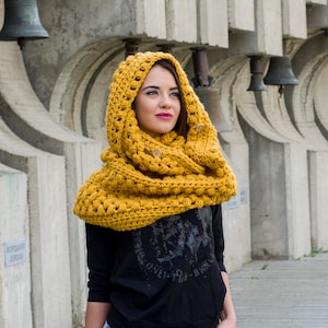 May include: A woman wearing a yellow crocheted cowl neck scarf with a black shirt and blue jeans. The scarf is thick and has a textured pattern. The woman is standing in front of a concrete wall with bells hanging from it.