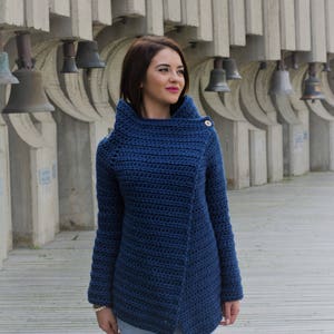 May include: A woman wearing a blue crocheted cardigan with a single button closure. The cardigan has a cowl neck and long sleeves. The woman is standing in front of a wall with bells.