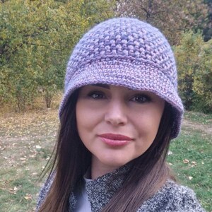 May include: A woman wearing a purple and pink crocheted hat with a wide brim. The hat is made with a textured stitch and has a soft, feminine look.