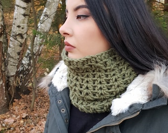 May include: A green crocheted neck warmer with a textured pattern. The neck warmer is worn by a person in a forest setting.