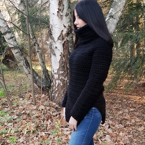 Puede incluir: Un suéter negro de cuello alto con mangas largas y un cuello alto. El suéter está tejido a crochet y tiene una longitud ligeramente más larga en la espalda. El suéter se usa con jeans azules.