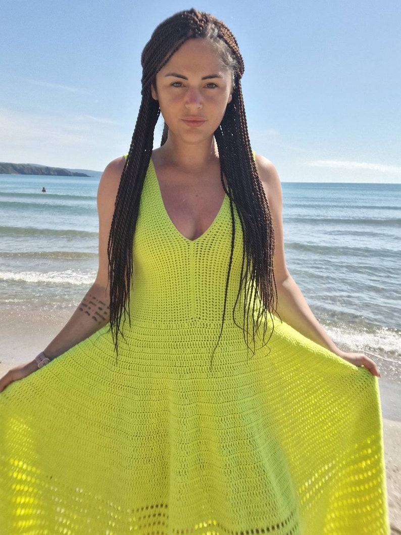 May include: A bright yellow crochet dress with a v-neckline. The dress is flowy and has a flared skirt. The model is wearing the dress on a beach.