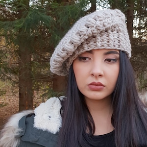 May include: A beige crocheted beret hat with a textured design. The hat is worn by a person with long black hair. The person is wearing a green jacket with a white fur trim.