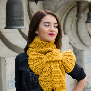 Puede incluir: Bufanda de ganchillo de color amarillo mostaza con un gran diseño de lazo. La bufanda está hecha de hilo grueso y se envuelve alrededor del cuello. El fondo presenta detalles arquitectónicos y campanas.
