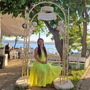 May include: A woman wearing a bright yellow crochet dress sits on a white metal bench under a white metal archway decorated with cherubs. The archway has a sign that reads "Welcome, enjoy your dinner!" in Cyrillic.