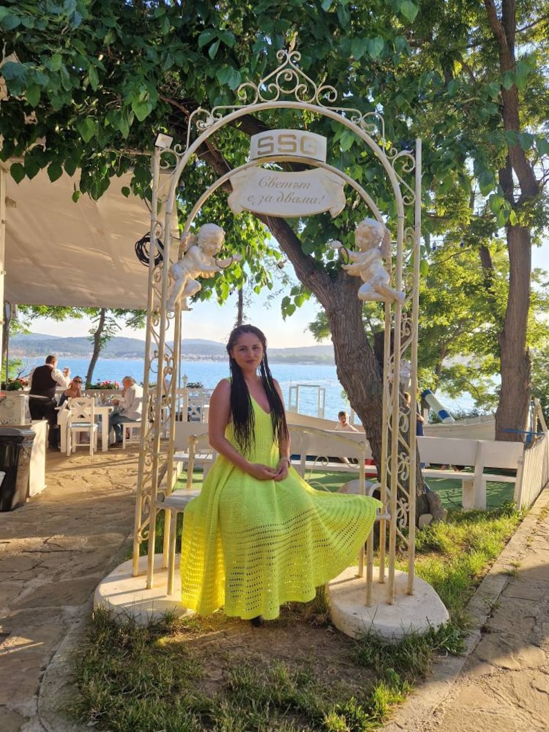 May include: A woman wearing a bright yellow crochet dress sits on a white metal bench under a white metal archway decorated with cherubs. The archway has a sign that reads "Welcome, enjoy your dinner!" in Cyrillic.