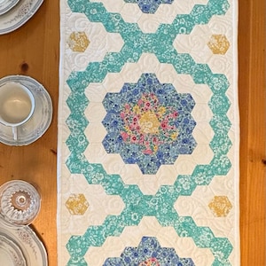 Pode incluir: Caminho de mesa acolchoado com um padrão floral repetido. O caminho de mesa apresenta um fundo branco com desenhos hexagonais florais turquesa, azuis e amarelos. O caminho de mesa está sobre uma mesa de madeira, com pratos e copos visíveis.