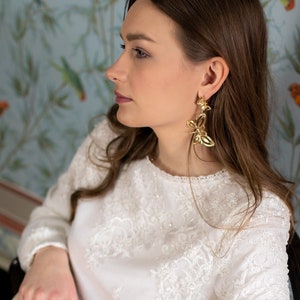 Op de afbeelding: Een vrouw draagt gouden vlinder oorbellen en een witte blouse met gedetailleerde kanten details. Ze houdt een crèmekleurige handtas met een geometrisch patroon vast. De achtergrond toont bloemenbehang.