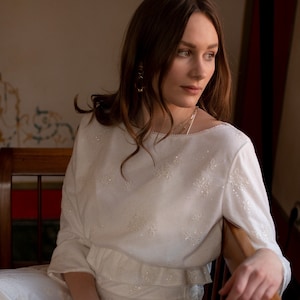 Op de afbeelding: Een wit, semi-transparant, lang mouwig top met een gerimpelde zoom en delicate bloemenborduursel. Het top wordt gedragen met een bijpassende witte rok.