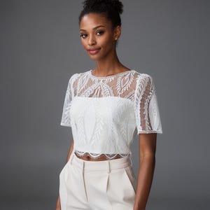 May include: A white short-sleeved top featuring an intricate lace overlay with a leaf and vine pattern. The garment has a round neckline and a scalloped hemline, layered over a solid white base fabric, paired with light beige high-waisted trousers.