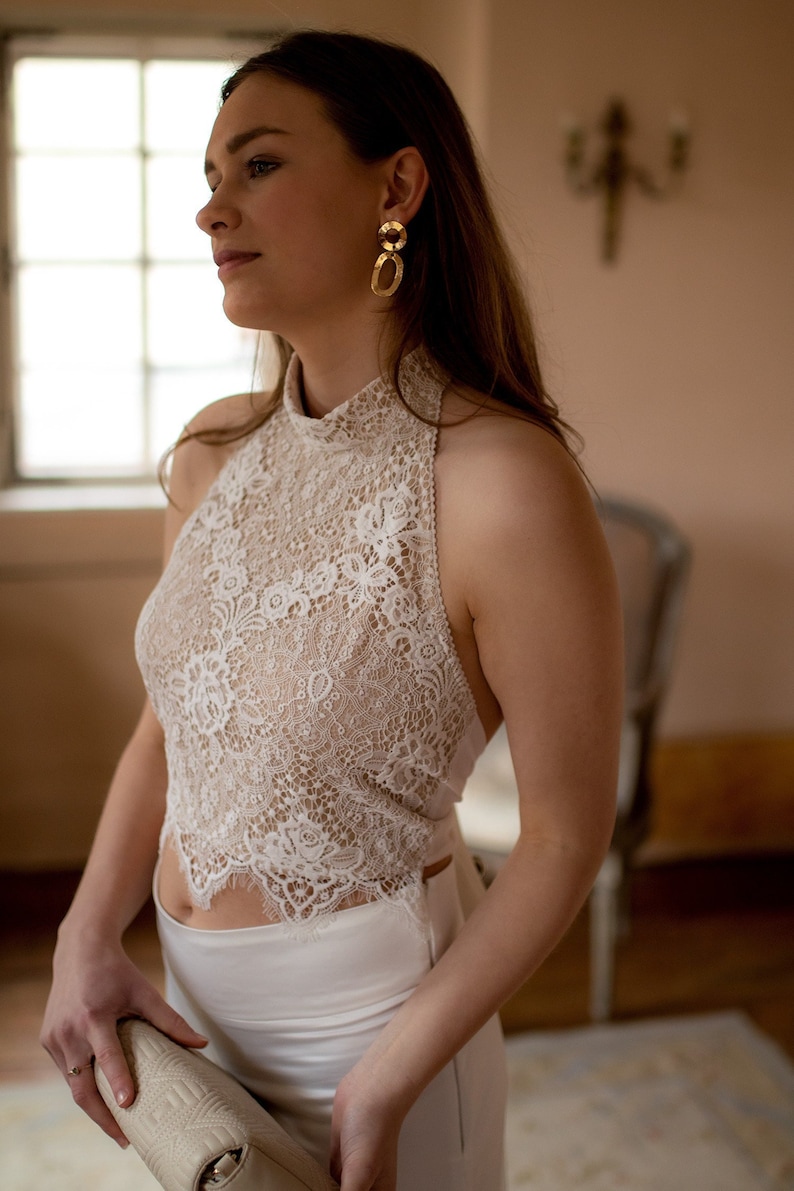May include: A woman wearing a white lace halter top with a high neckline and a white skirt. She is holding a white clutch purse and has gold hoop earrings.
