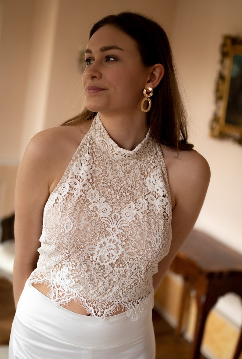 May include: A white lace halter top with a high neckline and a scalloped hem. The top is sheer and has a delicate floral pattern.