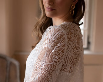 May include: A woman wearing a white lace bridal top with a delicate floral design. The top has a scoop neckline and three-quarter sleeves. She is also wearing a gold headband with small floral accents.