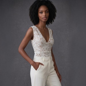 May include: A sleeveless ivory lace top with a deep V-neck, paired with matching ivory high-waisted pants. The lace top features a floral pattern. The model is standing in front of a gray background.