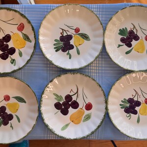 Peut inclure: Six assiettes en céramique blanches avec un bord festonné et une bordure verte. Chaque assiette présente un motif peint à la main de cerises, de raisins et d'une poire. Les assiettes sont disposées sur une surface à carreaux bleus et blancs.