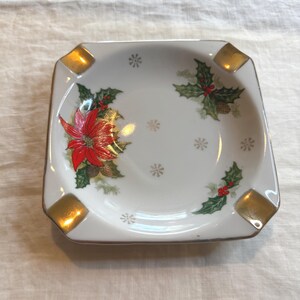 May include: Square white ashtray with gold accents and a Christmas theme. The ashtray features a red poinsettia, green holly leaves, red berries, and gold snowflakes. The corners have gold squares.