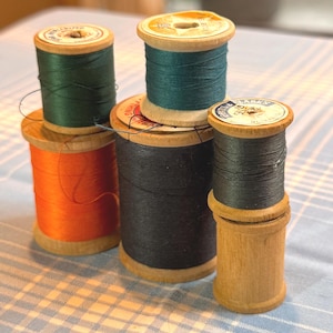 May include: A collection of vintage wooden spools of thread in various colors, including orange, green, and black. The spools are stacked and arranged on a light blue checkered surface. The thread is wound around the wooden spools.