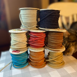 May include: Eight spools of leather cord in various colors, including beige, black, red, brown, orange, and blue. The spools are stacked in a pyramid shape on a white and gray checkered surface.