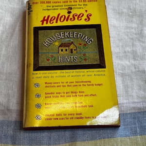May include: A yellow paperback book titled Heloise's Housekeeping Hints. The cover features a central illustration of a house in a frame with the text: Over 350,000 copies sold in the $3.95 edition, the greatest timesaver for the homemaker since TV dinners.