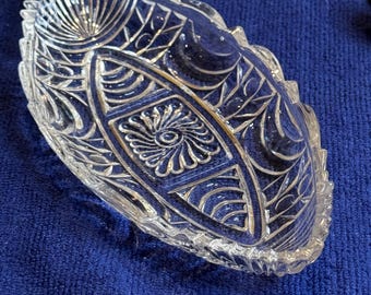 May include: Clear glass oval dish with intricate etched floral and geometric patterns. The dish has a scalloped edge and is set against a blue background. The design includes fan-like and spiral motifs.