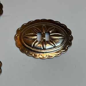 May include: Oval silver-toned metal buckle with a scalloped edge. The buckle has an embossed floral design and two rectangular slots in the centre. The metal has a slightly aged appearance.