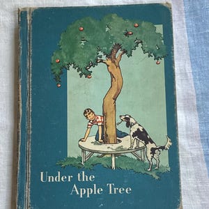 May include: A vintage hardcover book titled Under the Apple Tree with a teal cover. The illustration features a child and a black and white dog near a tree growing through a circular table. The book shows signs of wear along the edges and spine.