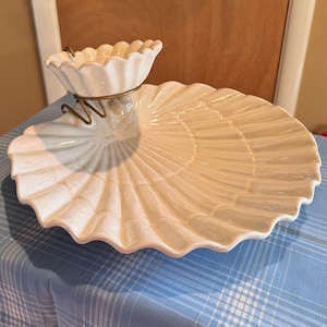Puede incluir: Un plato de cerámica blanco con forma de concha. El plato tiene un cuenco más pequeño y festoneado sujeto con un soporte de metal dorado. El plato grande con forma de concha tiene un diseño acanalado.