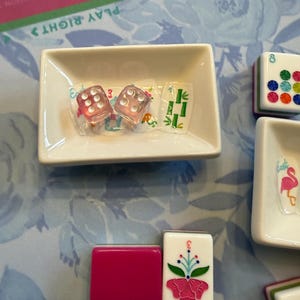 Może przedstawiać: Zbliżenie białej ceramicznej miski z dwiema r&oacute;żowymi kostkami i kafelkiem z motywem bambusa. Widocznych jest kilka kafelk&oacute;w do gry w Mahjonga, w tym jeden z r&oacute;żowym kwiatem i cyfrą 3.