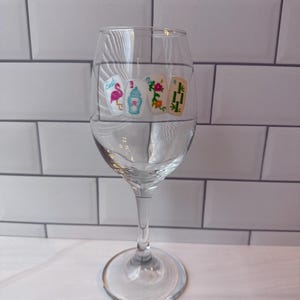 Pode incluir: Um copo de vinho de vidro transparente com um gr&aacute;fico impresso de quatro pe&ccedil;as de mahjong dispostas em fila. As pe&ccedil;as exibem um flamingo rosa, uma urna azul e vermelha, um padr&atilde;o floral e caules de bambu verdes com o texto East, 3, 2, 4.