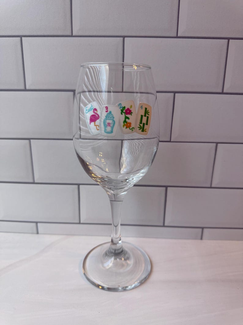 Pode incluir: Um copo de vinho de vidro transparente com um gr&aacute;fico impresso de quatro pe&ccedil;as de mahjong dispostas em fila. As pe&ccedil;as exibem um flamingo rosa, uma urna azul e vermelha, um padr&atilde;o floral e caules de bambu verdes com o texto East, 3, 2, 4.