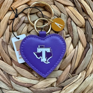 May include: A purple heart-shaped keychain with a gold ring and a small gold disc with the letter "A". The heart features a white and purple logo. The keychain is attached to a tag.