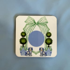 May include: Square white coaster with a blue circle in the center. The coaster features a green bow at the top, two topiary trees in blue and white vases, and two dogs. The background is a light blue.