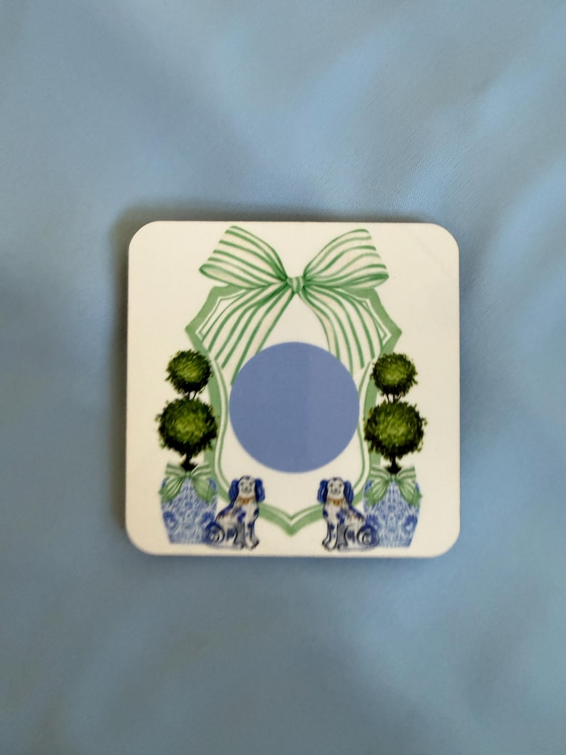 May include: Square white coaster with a blue circle in the center. The coaster features a green bow at the top, two topiary trees in blue and white vases, and two dogs. The background is a light blue.