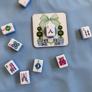 May include: A collection of white Mahjong tiles with blue and pink markings, scattered on a light blue surface. A decorative square tile features a green bow, topiary trees, and dog illustrations. The tiles display numbers, suits, and characters.