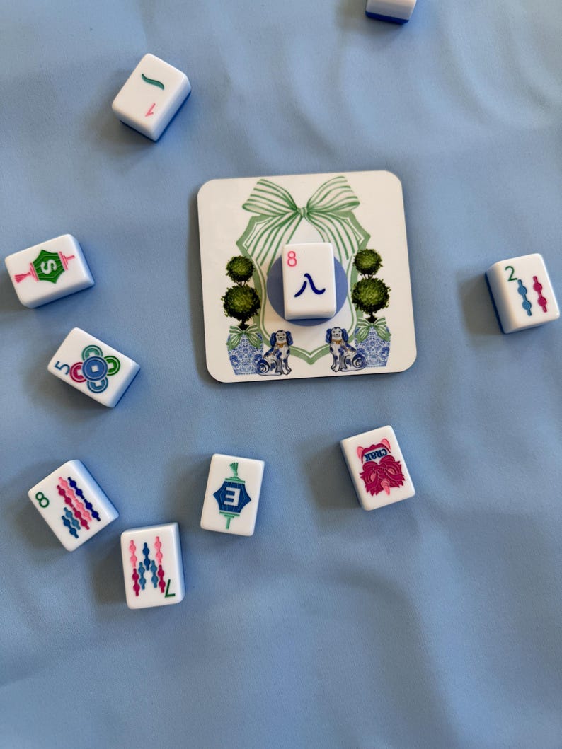 May include: A collection of white Mahjong tiles with blue and pink markings, scattered on a light blue surface. A decorative square tile features a green bow, topiary trees, and dog illustrations. The tiles display numbers, suits, and characters.