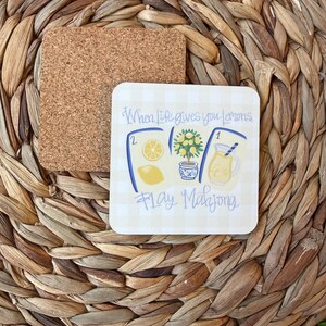 May include: A square coaster with a cork backing and a white surface featuring a light yellow gingham pattern. The design includes illustrations of a lemon, a lemon tree, and a pitcher of lemonade, with the text When Life gives you Lemons, Play Mahjong.