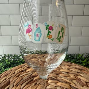 Pode incluir: Uma ta&ccedil;a de vinho de vidro transparente com quatro decalques retangulares brancos de pe&ccedil;as de mahjong. As pe&ccedil;as exibem um flamingo rosa com a palavra East, um pote de gengibre azul com o n&uacute;mero 3, uma trepadeira floral com o n&uacute;mero 2 e bambu verde com o n&uacute;mero 4.