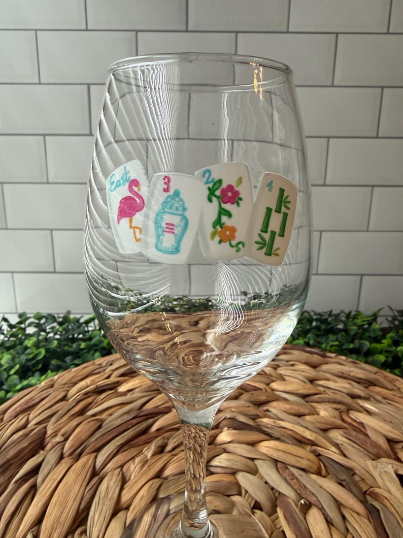 Pode incluir: Uma ta&ccedil;a de vinho de vidro transparente com quatro decalques retangulares brancos de pe&ccedil;as de mahjong. As pe&ccedil;as exibem um flamingo rosa com a palavra East, um pote de gengibre azul com o n&uacute;mero 3, uma trepadeira floral com o n&uacute;mero 2 e bambu verde com o n&uacute;mero 4.