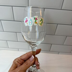 Pode incluir: Uma ta&ccedil;a de vinho de vidro transparente com um decalque de quatro pe&ccedil;as de mahjong dispostas em fila. As pe&ccedil;as exibem ilustra&ccedil;&otilde;es coloridas, incluindo um flamingo cor-de-rosa, uma urna azul, um padr&atilde;o floral e caules de bambu verdes.