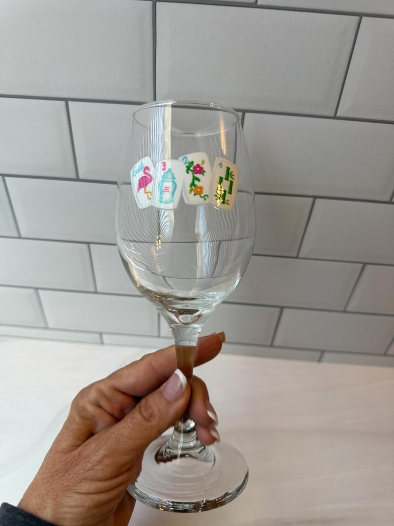 Pode incluir: Uma ta&ccedil;a de vinho de vidro transparente com um decalque de quatro pe&ccedil;as de mahjong dispostas em fila. As pe&ccedil;as exibem ilustra&ccedil;&otilde;es coloridas, incluindo um flamingo cor-de-rosa, uma urna azul, um padr&atilde;o floral e caules de bambu verdes.