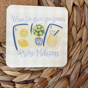 May include: A square coaster with a light yellow gingham pattern featuring three mahjong tiles. The tiles display a lemon slice, a lemon tree in a blue and white pot, and a pitcher of lemonade. The text reads, When life gives you lemons, play Mahjong.