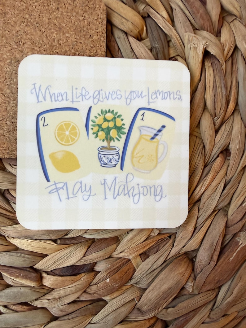 May include: A square coaster with a light yellow gingham pattern featuring three mahjong tiles. The tiles display a lemon slice, a lemon tree in a blue and white pot, and a pitcher of lemonade. The text reads, When life gives you lemons, play Mahjong.