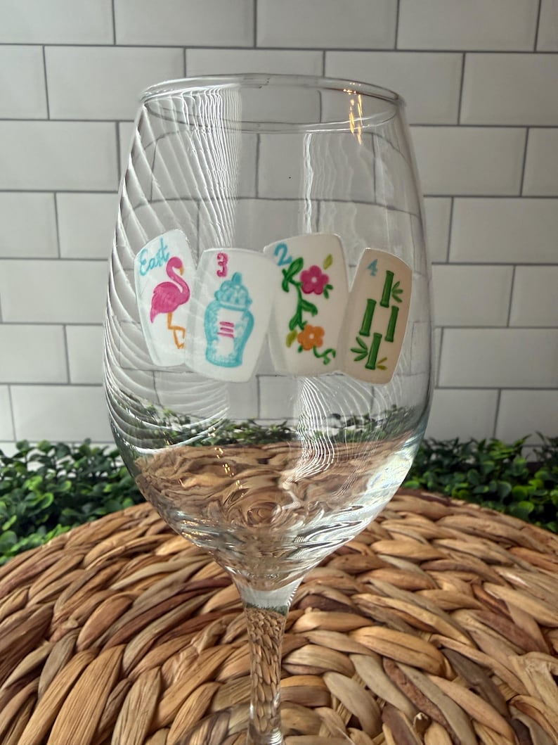 Pode incluir: Uma ta&ccedil;a de vinho de vidro transparente com quatro autocolantes coloridos de pe&ccedil;as de Mahjong. Os desenhos incluem um flamingo cor-de-rosa com a palavra East, um frasco de gengibre azul e branco com o n&uacute;mero 3, flores com o n&uacute;mero 2 e bambu verde com o n&uacute;mero 4.