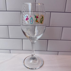 Pode incluir: Uma ta&ccedil;a de vinho de vidro transparente com um gr&aacute;fico impresso de quatro pe&ccedil;as de mahjong dispostas em fila. As pe&ccedil;as exibem ilustra&ccedil;&otilde;es coloridas, incluindo um flamingo rosa, uma urna azul, flores laranja e caules de bambu verdes.