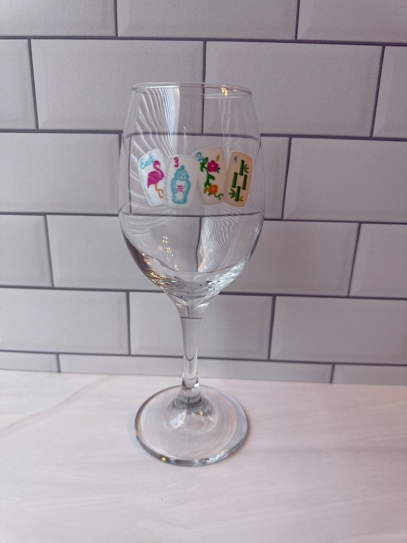 Pode incluir: Uma ta&ccedil;a de vinho de vidro transparente com um gr&aacute;fico impresso de quatro pe&ccedil;as de mahjong dispostas em fila. As pe&ccedil;as exibem ilustra&ccedil;&otilde;es coloridas, incluindo um flamingo rosa, uma urna azul, flores laranja e caules de bambu verdes.
